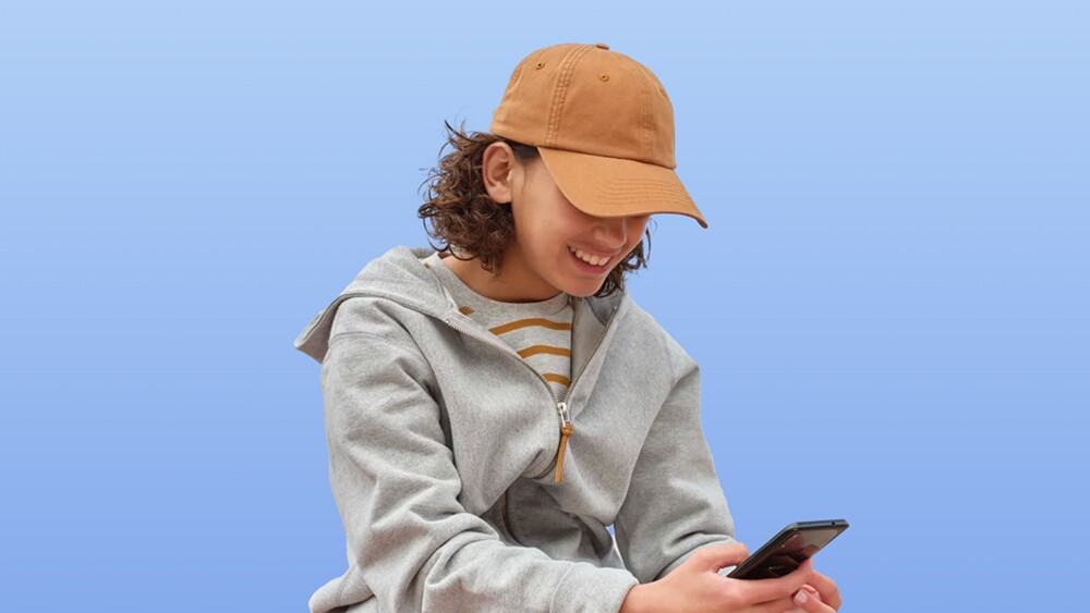 A smiling teenager in a tan baseball cap, gray zip-up hoodie, and striped shirt looks down at their phone while standing against a bright blue background, representing a satisfied Buy with Prime customer.
